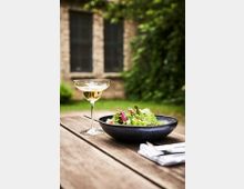 Eine Holzbank im Freien mit einem Glas Sekt und einem Teller mit frischem Salat. Im Hintergrund ein Gebäude aus Backstein mit Fenstern, umgeben von grüner Vegetation.