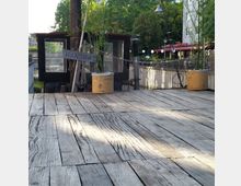 Holzdeck eines schwimmenden Pontons mit Pflanzenkübeln und einer kleinen Kabine, beschriftet mit „Hanseat“. Im Hintergrund sind Bäume, Lampen und ein Gebäude mit einem Café zu sehen.