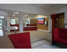 Empfangsbereich eines modernen Gebäudes mit roten Sitzmöbeln, einem Empfangstresen aus Holz und Glas sowie einer digitalen Anzeigetafel an der Wand. Im Hintergrund sind große Fenster und eine Glastür, die Zugang zu einem Außenbereich bieten.