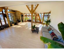 Wohnzimmer mit hellem Laminatboden, großen bodentiefen Fenstern mit Holzrahmen und Holzbalken. Links ein Kamin aus Sichtstein, rechts eine großzügige Couch mit bunten Kissen, umgeben von zahlreichen Grünpflanzen.