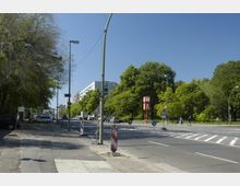 Straßenszene in einer urbanen Umgebung mit breitem Gehweg links, einem Streifen von Bäumen rechts und Wohngebäuden im Hintergrund. Verkehrsschilder, eine Fußgängerampel sowie eine rote Uhrsäule sind entlang der Straße sichtbar.