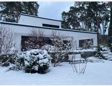 Modernes, weißes Flachdachhaus mit klaren, rechteckigen Formen und großer überdachter Terrasse, umgeben von hohen Kiefern. Im Vordergrund liegt ein verschneiter Garten mit Sträuchern, niedriger Steinmauer und einer Bank im Schnee.
