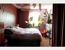 Schlafzimmer mit rosafarbenen Wänden und einem großen Bett, das mit einer flauschigen, pastellrosa Decke bedeckt ist. Der Raum ist mit Pflanzen dekoriert, hat einen Holzboden, einen orangenen Stuhl sowie Vorhänge in einem kräftigen Rotton, die das Fenster teilweise verdecken.