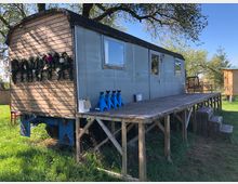 Ein renovierter Bauwagen mit einer Holzveranda, aufgeständert über einer Wiese. Die Vorderseite des Wagens ist mit Holz verkleidet und mit Blumendekorationen versehen, während die Seitenwände aus Metall bestehen. Im Hintergrund sind Bäume und ein weiteres kleines Holzgebäude sichtbar.
