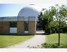 Kuppelförmiges Gebäude mit einer Verkleidung aus weißen Platten, daneben ein einstöckiger, mit rotbraunen Ziegeln verkleideter Anbau mit Fenstern. Im Vordergrund führt ein gepflasterter Weg durch eine Grünanlage mit Bäumen und Sträuchern.