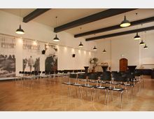 Ein moderner Veranstaltungsraum mit hellem Holzboden, schwarzen Stühlen in Reihen, Hängelampen an der Decke und schwarz-weißen Wandbildern. Im Hintergrund sind Stehtische, eine Pflanze sowie eine geschlossene Holztür sichtbar.