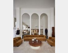 Ein stilvolles Modegeschäft mit einer eleganten Inneneinrichtung, bestehend aus beige gepolsterten Sofas, einem runden Holztisch und weißen Wänden mit dekorativen Rundbögen. Im Hintergrund stehen Schaufensterpuppen mit Kleidung sowie offene Regale mit weiteren Kleidungsstücken, während große Fenster viel Tageslicht hereinlassen.