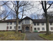 Zweistöckiges Gebäude mit weißer Fassade, dunklem Satteldach und einem zentralen, leicht vorspringenden Eingangsbereich mit Rundbögen und großen Fenstern. Im Vordergrund stehen kahle Bäume, und der Boden ist mit Gras und einem gepflasterten Weg gestaltet.