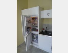 Ein offener, weißer Einbaukühlschrank mit mehreren Fächern, gefüllt mit Lebensmitteln und Getränken, in einer modernen Küche mit gelber Wand und grauem Boden. Rechts daneben befindet sich eine Arbeitsplatte mit einer Flasche und einem Glas.