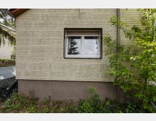 Außenwand eines Wohnhauses mit hellen Klinkersteinen und einem zentralen Doppelfenster mit Rollladen. Unten ist ein dunkler Sockelputz zu sehen; davor wachsen Sträucher und Gartenpflanzen.
