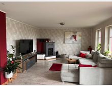 Wohnzimmer mit Ecksofa, Holzfußboden und roten Akzenten. Im Raum befinden sich ein Flachbildfernseher, eine dekorative Feuerstelle, ein kleiner Holztisch und mehrere Pflanzen. Drei große Fenster rechts lassen viel Tageslicht ein und beleuchten die hellgraue Tapete mit floralem Muster.