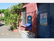 Außenansicht eines niedrigen Backsteingebäudes mit großem Fenster und blauer Tür, an der „Theater im Bergmannkiez“ steht. Vor der Fassade rankt eine blühende Kletterpflanze, daneben befindet sich eine Informationstafel und ein Plakat, entlang eines gepflasterten Hofwegs.