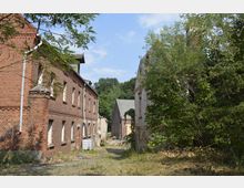 Ehemaliges Fabrik- oder Lagergelände mit roten Ziegelgebäuden, von denen einige Fenster zugemauert sind. Die Umgebung ist verwildert mit hohem Gras und Bäumen, im Hintergrund sind weitere beschädigte Gebäude sichtbar.