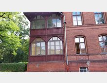 Backsteinfassade eines historischen Gebäudes mit einem aus Holz gestalteten Erker. Die Fassade ist rotbraun, verziert mit dekorativen Holzelementen und großen Sprossenfenstern. Im Hintergrund sind Bäume und Sträucher sichtbar.