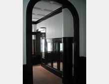 Ein historischer Innenraum mit dunklen Holzpaneelen an den Wänden, einem gewölbten Türrahmen und einem Teppichboden. Durch Fenster dringt Tageslicht in den Raum, und die Decke ist mit rechteckigen Vertiefungen verziert.