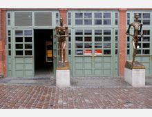 Außenansicht eines Backsteingebäudes mit grünen Fenstern und Türen, flankiert von zwei bronzenen, schlanken Skulpturen auf Betonsockeln; der gepflasterte Boden führt zu einem offenen Eingang, der einen Blick ins Gebäudeinnere gewährt.