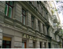 Fassade eines alten Wohnhauses mit verwittertem Putz, hohen Fenstern mit Holzrahmen und dezenten Stuckverzierungen, daneben weitere historische Gebäude mit Balkonen und Dekorelementen. Im unteren Bereich ist ein verblasstes Schild mit dem Wort "FUS(S)..." erkennbar.