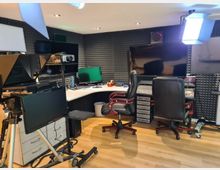 Ein Studio mit verschiedenen Bildschirmen, Aufnahme- und Kontrollgeräten sowie zwei schwarzen Bürostühlen vor einer U-förmigen Arbeitsfläche. Wände sind mit schallabsorbierenden Schaumstoffplatten verkleidet, und Studiobeleuchtung sowie Kameraausrüstung sind sichtbar.