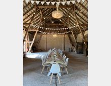 Großer, rustikaler Scheunenraum mit Holzbalken und hoher Decke. Ein langer, gedeckter Holztisch mit weißen Stühlen bildet den Mittelpunkt, dekoriert mit Blumen und Gläsern; darüber hängen ein großer Kronleuchter und weiße Wimpelketten.