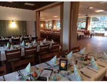 Ein großer, geschmackvoll dekorierter Veranstaltungsraum mit gedeckten Tischen, die mit weißen Tischdecken, bedruckten Servietten und Kerzendekorationen geschmückt sind. Der Raum ist in mehrere Bereiche unterteilt, hat Holzmöbel, große Fenster mit Blick ins Freie und eine warme Beleuchtung.