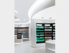 Moderne Apotheke mit weißen, geschwungenen Wänden und Regalen, gefüllt mit medizinischen und kosmetischen Produkten in geordneten Reihen. Die Umgebung ist hell beleuchtet, mit einem minimalistischen und sauberen Design.