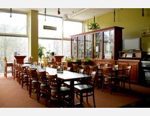 Ein gemütlicher Innenraum eines Restaurants oder Cafés mit Holzstühlen und -tischen, großen Fenstern, die Tageslicht hereinstrahlen, und einer Vitrine im Hintergrund mit Pokalen und Dekorationen. Pflanzen und eine helle Wandgestaltung ergänzen das Ambiente.