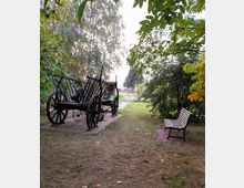 Historischer Holzwagen mit Holzspeichenrädern auf einem gepflasterten Weg in einer parkähnlichen, grünen Umgebung mit Bäumen; daneben steht eine hölzerne Sitzbank. Im Hintergrund führt ein Weg zu einem Zaun.