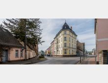 Straßenszene in einer Kleinstadt mit einer gelben, mehrstöckigen Eckvilla mit spitzem Dach im Zentrum des Bildes. Umgeben von weiteren Wohnhäusern, schmalen Gehwegen, einigen parkenden Autos und einem Baum im Vordergrund.