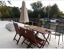 Holzterrasse auf einem Boot mit einem Tisch, Holzstühlen und einem Sonnenschirm, die neben einem ruhig gelegenen Fluss mit angedockten Booten stehen; im Hintergrund dichter Baumbestand.