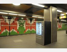 U-Bahn-Station mit moderner Wandgestaltung aus roten, grünen und weißen Fliesen, die den Schriftzug „Mierendorffplatz“ tragen. Im Vordergrund befindet sich ein beleuchteter Snackautomat, darüber eine Decke mit Beleuchtungselementen.