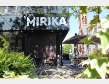 Außenbereich eines modernen Cafés mit der Aufschrift "MIRIKA" an der Fassade. Es gibt Tische und Stühle auf einem mit Kopfsteinpflaster ausgelegten Platz, umgeben von grünen Sträuchern und Bäumen; im Hintergrund ist ein Kirchturm sichtbar.