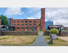 Backsteinbau mit einem hohen Turm im Zentrum, flankiert von modernen Glasfassaden. Im Vordergrund ein gepflasterter Weg, eine Trockenmauer und bepflanzte Holzkästen in einer Wiesenlandschaft.