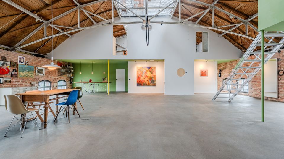 Großer, loftartiger Innenraum in einer umgebauten Industriehalle mit sichtbaren Stahlträgern, hohem Holztonnen­dach und Backsteinwänden. Offene Fläche mit Esstisch und Stühlen, kleiner Küchenzeile, Wandkunst sowie einer Metalltreppe zu einer oberen Galerieebene.