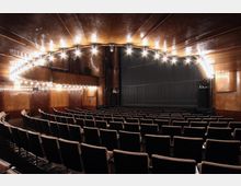 Innenansicht eines Theatersaals mit leicht gebogenem Parkett, Reihen von gepolsterten Sitzen in Braun, einer wellenförmigen Decke mit eingebauten Lichtern und einer Bühne mit geschlossenem, geripptem Vorhang. Die Wandverkleidungen sind aus dunklem Holz.