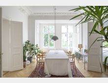 Lichtdurchfluteter Raum mit hohen Decken, verziertem Stuck und großen weißen Sprossenfenstern. Mittig steht ein Tisch mit weißer Tischdecke auf einem Teppich, umgeben von Holzstühlen; Pflanzen und schlichte Möbel ergänzen die Einrichtung.