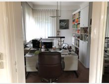 Ein Bürozimmer mit einem Schreibtisch in der Mitte, umgeben von mehreren vollen Bücherregalen an der rechten Wand. Auf dem Schreibtisch befinden sich Computerbildschirme, Büromaterial und Unterlagen, daneben steht ein dunkler Bürostuhl. Ein großes Fenster mit weißen Vorhängen sorgt für Tageslicht.