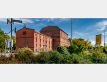 Backstein-Gebäude im historischen Industriestil, umgeben von Vegetation und Straßenbeschilderungen. Im Hintergrund sind Bäume unter einem klaren blauen Himmel zu sehen.