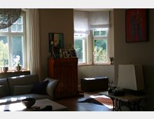 Wohnzimmer mit großen Fenstern und viel Tageslicht. Im Raum befinden sich eine helle Couch, ein Holztisch, eine antike Kommode mit gerahmten Fotos und ein alter Truhe, umgeben von schlichten Vorhängen und dekorativen Accessoires.