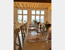 Langer, festlich gedeckter Holztisch in einem Raum mit weiß gestrichenen Wänden, großen Sprossenfenstern und einer Decke mit sichtbaren Holzbalken. Im Hintergrund ist durch die Fenster eine Aussicht auf eine Landschaft zu erkennen.