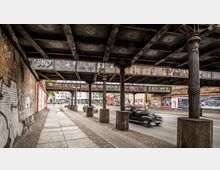 Straßenansicht unter einer alten Eisenbahnbrücke mit rostigen Stahlträgern und verzierten Gusseisenstützen. Auf einer Seite befindet sich ein Fußweg neben einer Backsteinmauer mit Graffiti, während Autos auf der Fahrbahn unter der Brücke vorbeifahren; im Hintergrund sind Gebäude und Werbetafeln zu sehen.