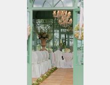 Ein dekorierter Innenraum eines Pavillons mit grünen Metall- und Glaselementen. Der Raum ist für eine Feierlichkeit eingerichtet, mit weiß überzogenen Stühlen, einem Tisch mit Blumenarrangements und einem Kronleuchter an der Decke.