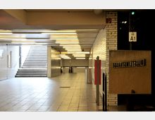 Unterirdischer U-Bahn-Bereich mit weißen Fliesenwänden, hell beleuchteter Decke und Bodenfliesen. Treppen führen nach oben zu einem Ausgang, und Wegweiser mit Beschriftungen sind sichtbar; ein Spiegel zeigt den Schriftzug „Ullsteinstraße“ spiegelverkehrt.