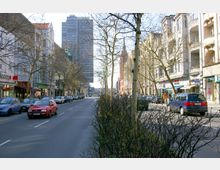 Städtische Straßenansicht mit mehrstöckigen Wohn- und Geschäftshäusern auf beiden Seiten, parkenden und fahrenden Autos sowie kahlen Bäumen entlang eines Mittelstreifens. Im Hintergrund ist ein moderner Hochhauskomplex zu sehen.