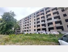 Ein leerstehendes, mehrstöckiges Gebäude aus Beton mit zahlreichen offenen Fensteröffnungen und Graffiti an den Wänden, umgeben von wilder Vegetation und einzelnen Bäumen. Rechts im Vordergrund steht ein geparktes Auto.