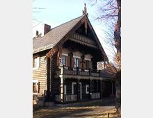 Holzhaus im traditionellen Stil mit Satteldach, verzierten Holzverkleidungen und einem Balkon. Es steht in einer ländlichen Umgebung mit kahlen Bäumen und vereinzeltem Sonnenlicht im Vordergrund.