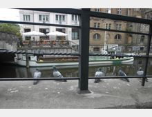 Blick auf einen kleinen Kanal mit einer weißen Ausflugsboot-Anlegestelle im Vordergrund, mehreren Tauben auf einem Geländer und einem Gebäude mit Restaurant und Terrassenbereich im Hintergrund. Rechtsseitig ist eine alte Backsteinfassade zu sehen.