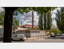 Straßenszene mit einem großen Baum im Vordergrund, einem Industriegebäude mit rotem Ziegeldach und einem hohen Schornstein im Hintergrund. Umgeben von hohen Bäumen und geparkten Autos, befindet sich ein flaches Gebäude an der Straßenecke.