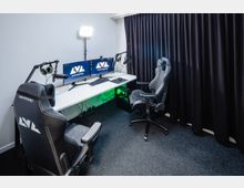 Ein moderner Gaming- und Streaming-Raum mit zwei Gaming-Stühlen, einem weißen Schreibtisch, zwei Monitoren, einem Mikrofon, einem Lichtpanel und einem PC mit grüner Beleuchtung. Im Hintergrund befinden sich dunkle Vorhänge vor einer weißen Wand.
