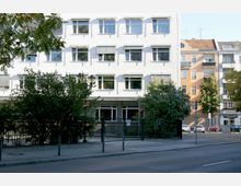 Ein weißes, mehrstöckiges Gebäude mit rechteckigen Fenstern und heruntergelassenen Jalousien, umgeben von Bäumen und Sträuchern im Vordergrund. Vor dem Gebäude verläuft ein Gehweg mit einer schwarzen Metallzaunanlage.