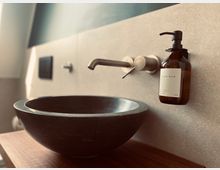 Waschbecken in moderner Gestaltung mit einer dunklen Steinschale, einer minimalistischen Wandarmatur aus Metall und einer Wandhalterung mit Spenderflasche für Handseife; Hintergrund aus hellen Wandfliesen.